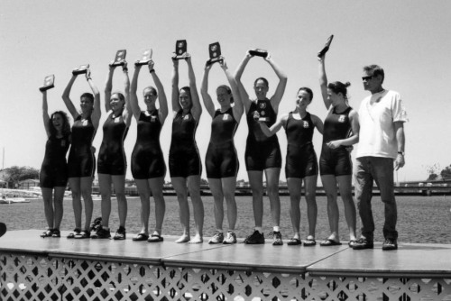 A black and white photo of a row of female athletes holding up plaques next to their coach on a dock.