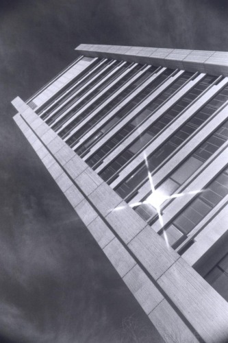 A black and white photograph of the Sciences Library, a tall, brutalist building shot from a ground angle so that the building stretches towards the sky, with one of the windows catching the shine of the sun.