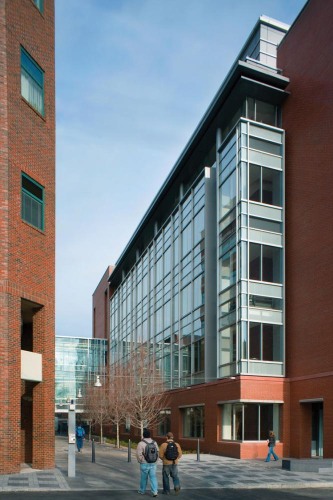Two students walk towards a path between two buildings made from brick and glass.