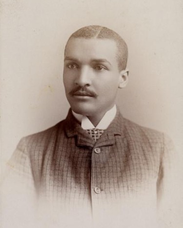 A black and white photo of a black man with a short haircut in a suit with a check pattern making a serious expression with a soft vignette around him