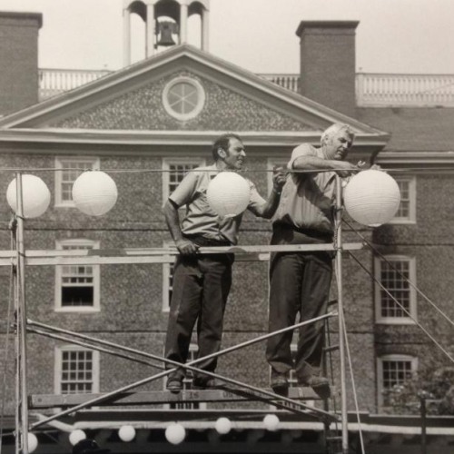 A black and white photograph of two workers standing on scaffolding in front of a university building hanging paper lanterns above the green.