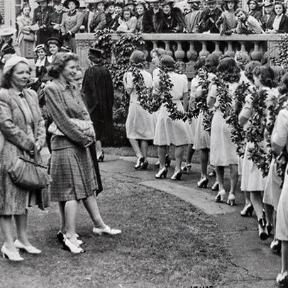 A black and white photograph of female students in traditional dress looking on as a procession of female students dressed in white with ivy sashes walk through Pembroke campus.