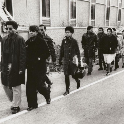 A black and white photograph of a line of Black students walking on the street with serious expressions and dressed for winter weather.