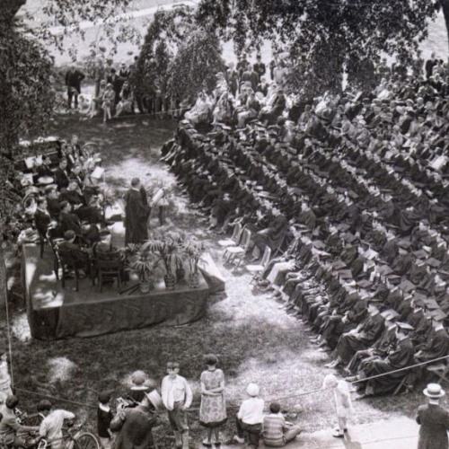 A black and white photograph of rows of students wearing caps and gowns sitting before onlookers and speakers.