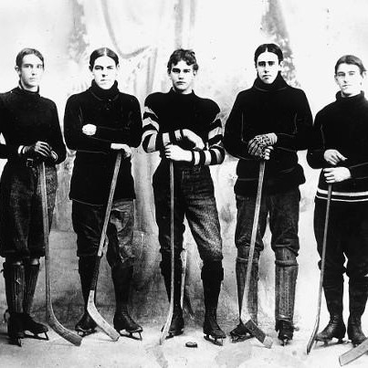 A black and white photograph of seven men in hockey uniforms standing in a line with hockey sticks
