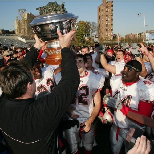 A coach holds a large trophy over the heads of football players in uniform.