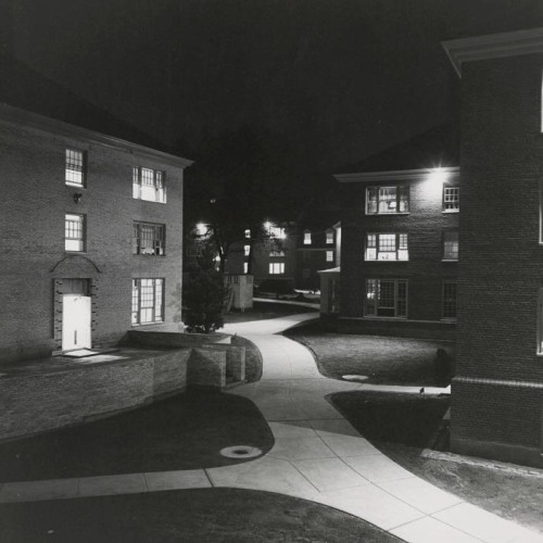 A black and white nighttime photograph of a few brightly lit dormitory buildings and a path through them.