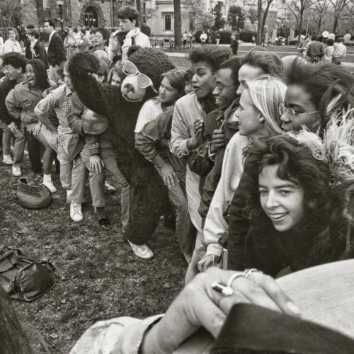 A row of students sitting on each other in a line, with Bruno, Brown's bear mascot, sitting in the center with sunglasses falling off his face.