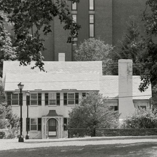 A black and white photo of a small white building with dark gables in front of a larger brick building.