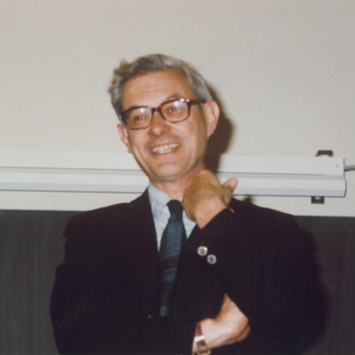 A color photograph of an older white man in glasses and a suit and tie smiling and adjusting his collar while the other arm is crossed.