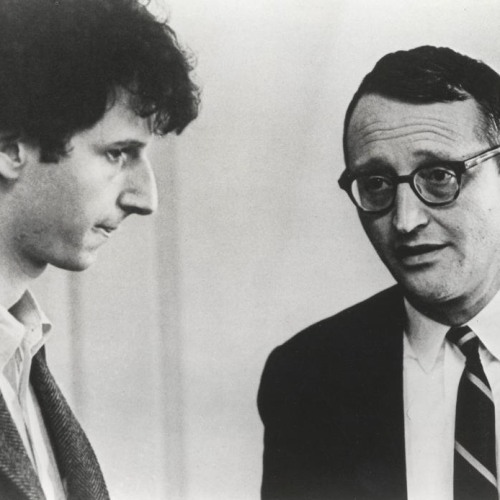 A black and white photograph of two men, one is younger facing profile and looking down, and the other is older looking at the younger man, who is a student.