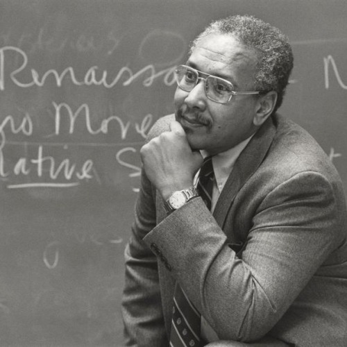 A black and white photo of an older Black man in a suit and tie resting his head on his hand in a thinking position in front of a chalk board with writing on it.