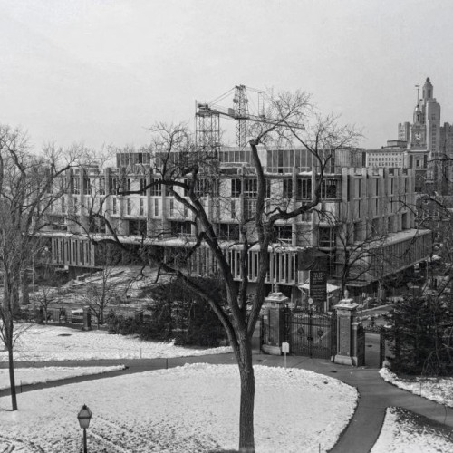 A black and white photograph of a large brutalist building under construction surrounded by snowy patches of grass, cleared paths, and wintry trees.